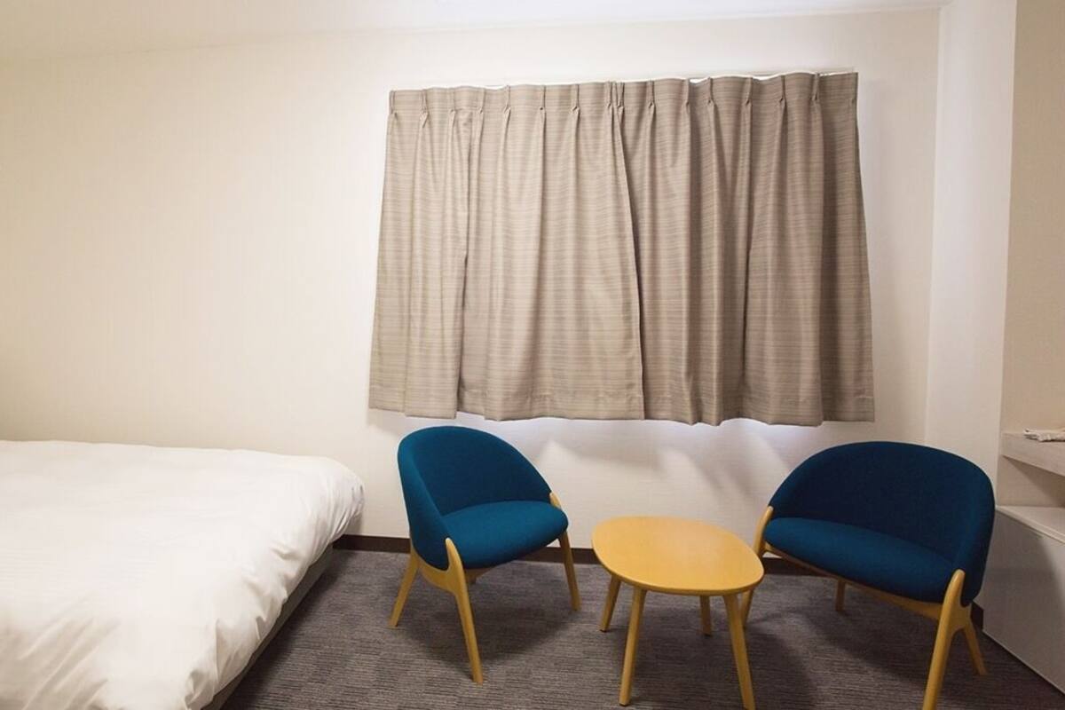 Une chambre d’hôtel avec un lit, deux chaises bleues, une petite table ronde et une fenêtre avec des rideaux.