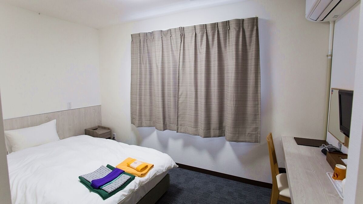 Een hotelkamer met een bed, een bureau met een computer, een stoel, een nachtkastje en een raam met gordijnen.