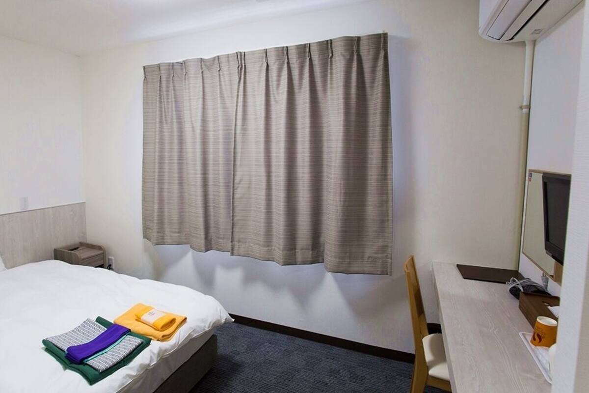 Une chambre d’hôtel comprenant un lit, un bureau avec un ordinateur, une chaise, une table de chevet et une fenêtre avec des rideaux.