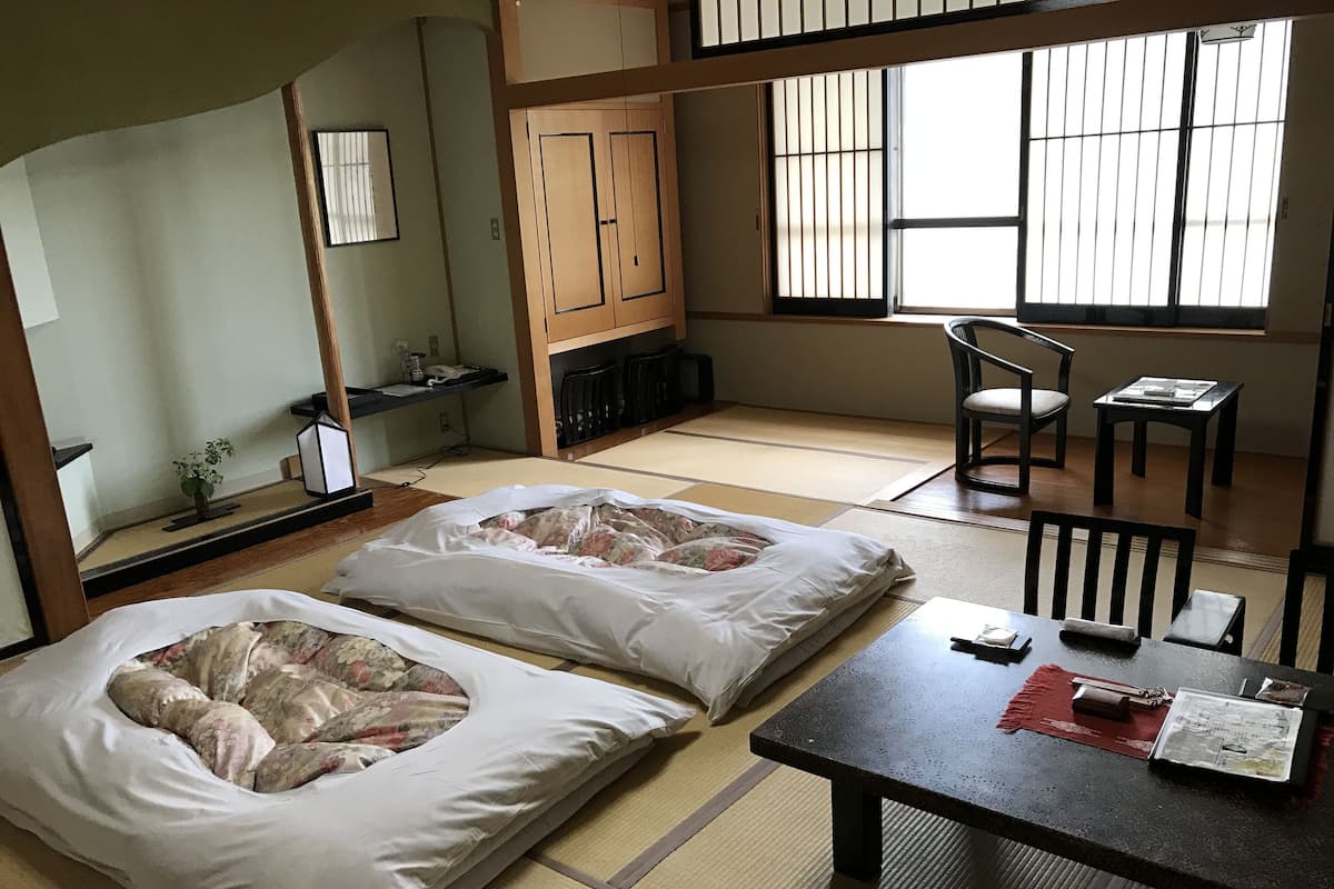 A traditional Japanese room with tatami flooring, two tatami mats with floral bedding, a wooden table with a newspaper, and a sliding door with a view.