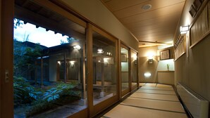 A corridor with sliding glass doors leading to a garden area, wooden flooring, and wall-mounted lighting. - Kazennosyo (Kuroishi)