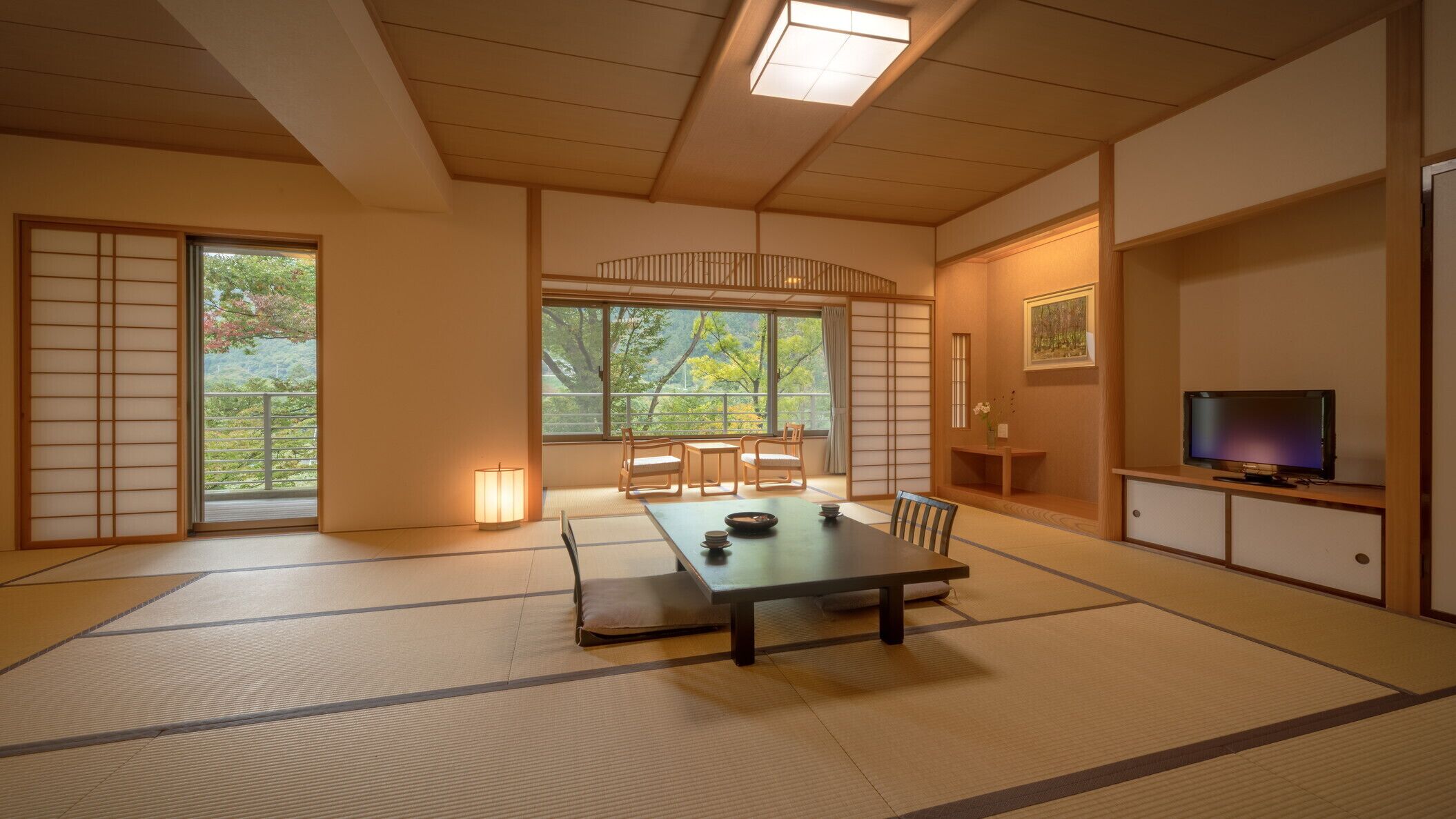 A traditional Japanese room with tatami flooring, a low table with cushions, a television, and a view of trees through large windows.
