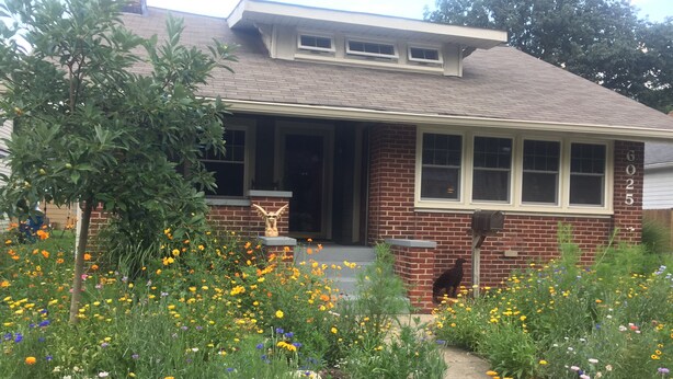 Une maison en briques avec un porche à l’avant, une boîte aux lettres et un jardin où poussent de nombreuses fleurs aux couleurs vives.