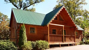 A log cabin with a green metal roof, a porch with a railing, and a small window.