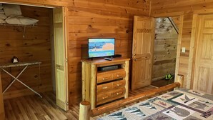 A wooden cabin room with a bed, a TV on a wooden stand, a small table, and a door leading to another room.