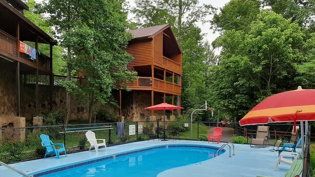A swimming pool with a wooden deck, surrounded by greenery and a building with a balcony.