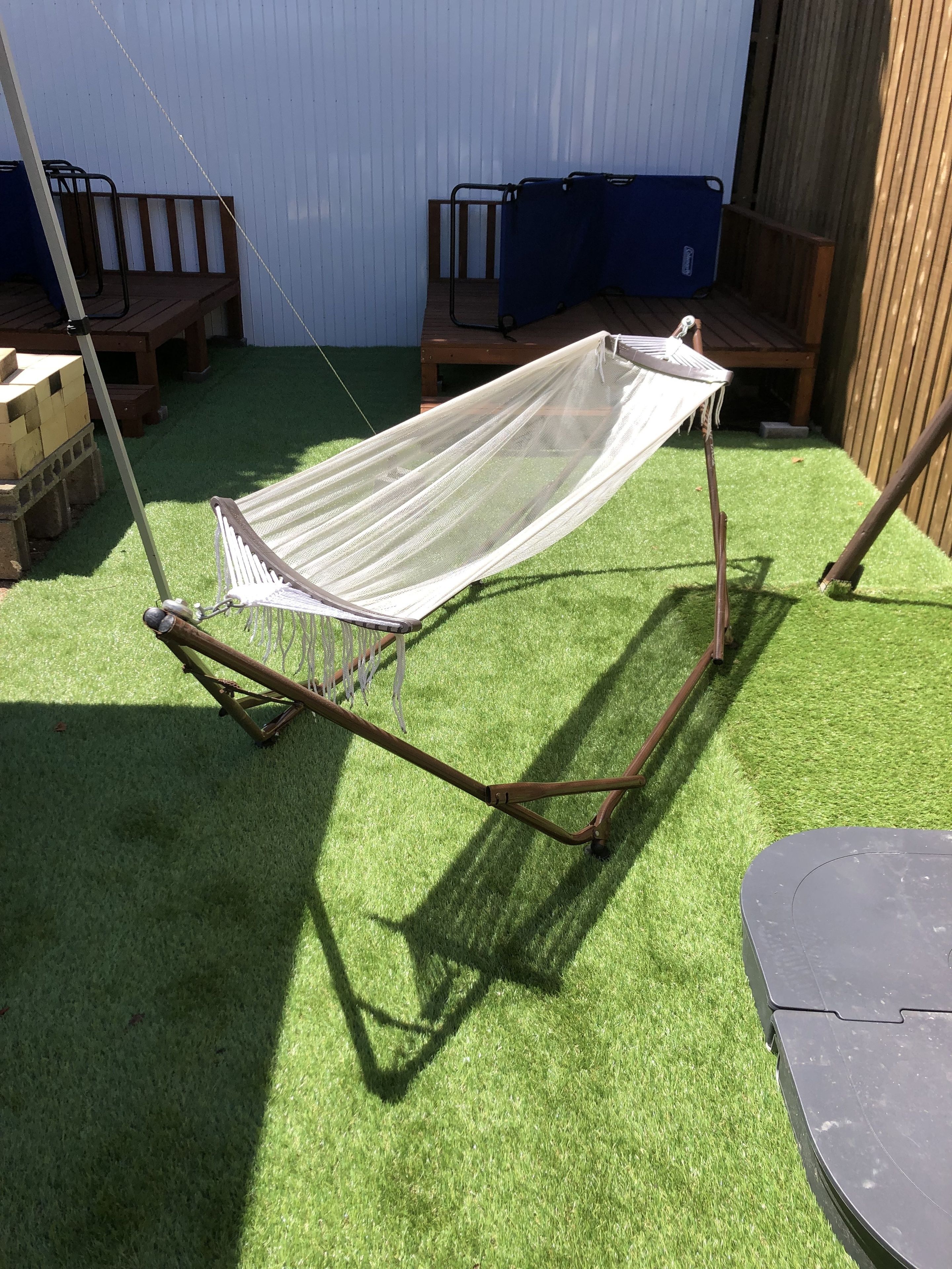 A hammock with a white net, wooden frame, and metal support rods, set on artificial grass in an outdoor area with wooden benches and a table.