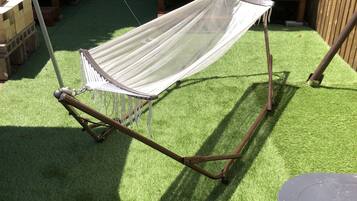 A hammock with a white net, wooden frame, and metal support rods, set on artificial grass in an outdoor area with wooden benches and a table.