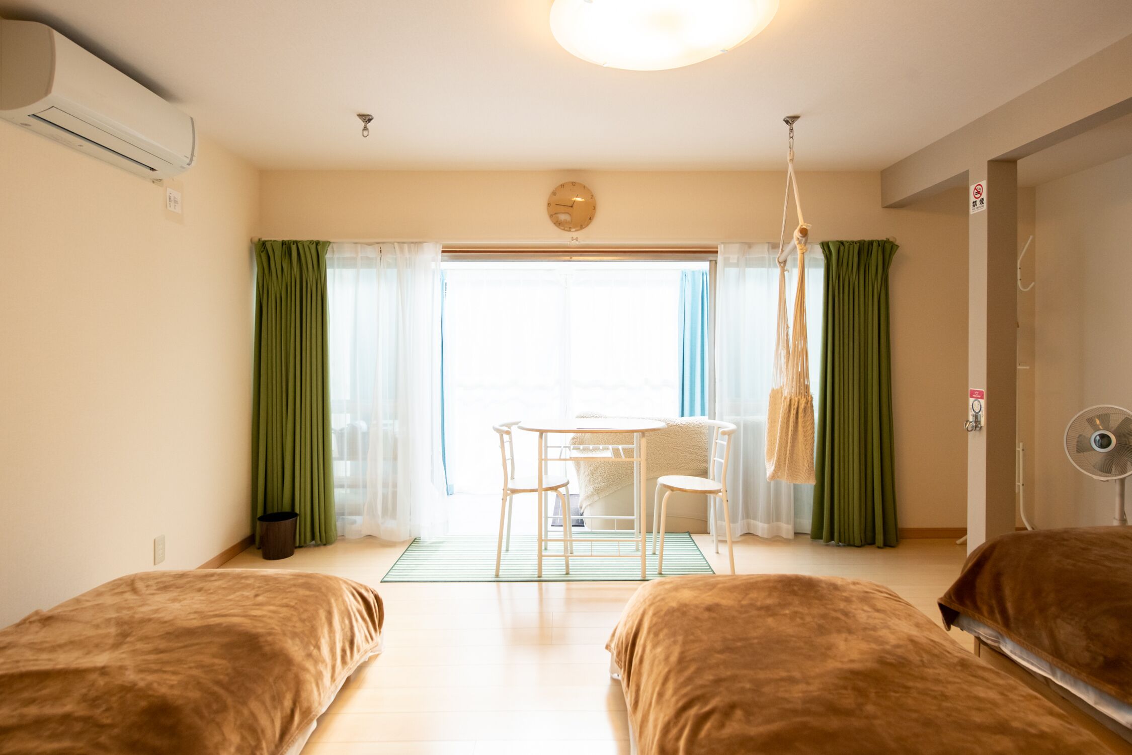 A hotel room with two beds, a dining table with chairs, a hanging light fixture, and a view of the outdoors through a large window.