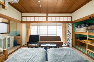 Una habitación tradicional japonesa con tatamis, un futón, una mesita, una silla, una pequeña cocina y un armario con ropa colgada.