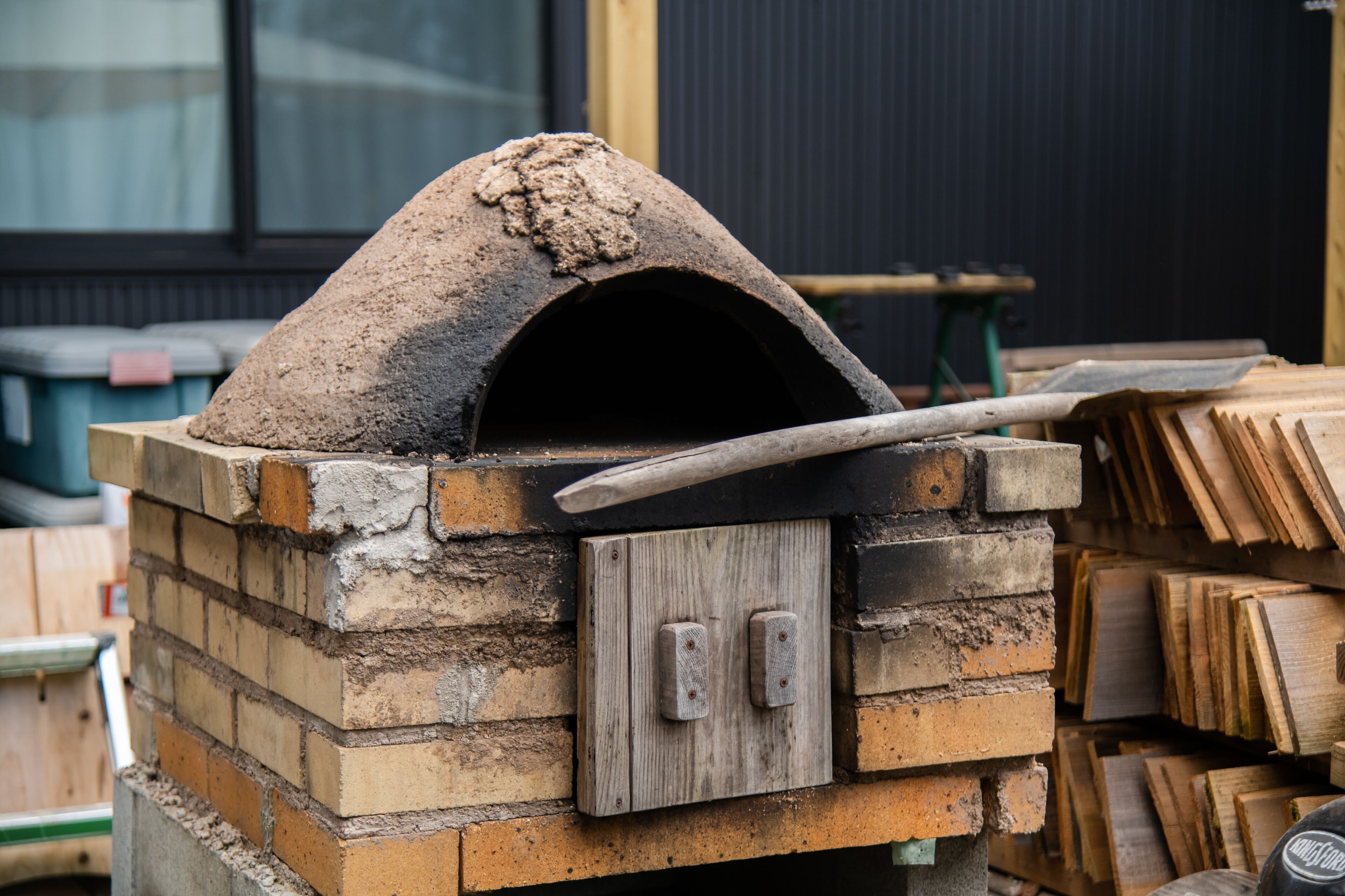 Un four extérieur en briques, doté d’une poignée en bois, installé devant un décor composé de bois de chauffage empilé et d’une fenêtre.