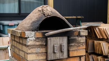 Un four extérieur en briques, doté d’une poignée en bois, installé devant un décor composé de bois de chauffage empilé et d’une fenêtre.