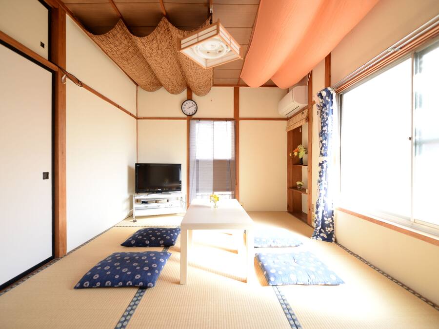 Una habitación tradicional japonesa con suelo de tatami, un televisor, una mesita con un florero y un ventanal con cortina.