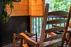 A wooden chair with a cushioned backrest and armrests, positioned on a wooden deck with a view of greenery outside.