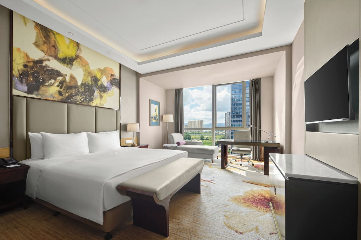 Ein Hotelzimmer mit einem großen Bett, einem Schreibtisch, einem Sessel, einem Fernseher und Blick auf die Stadtlandschaft.