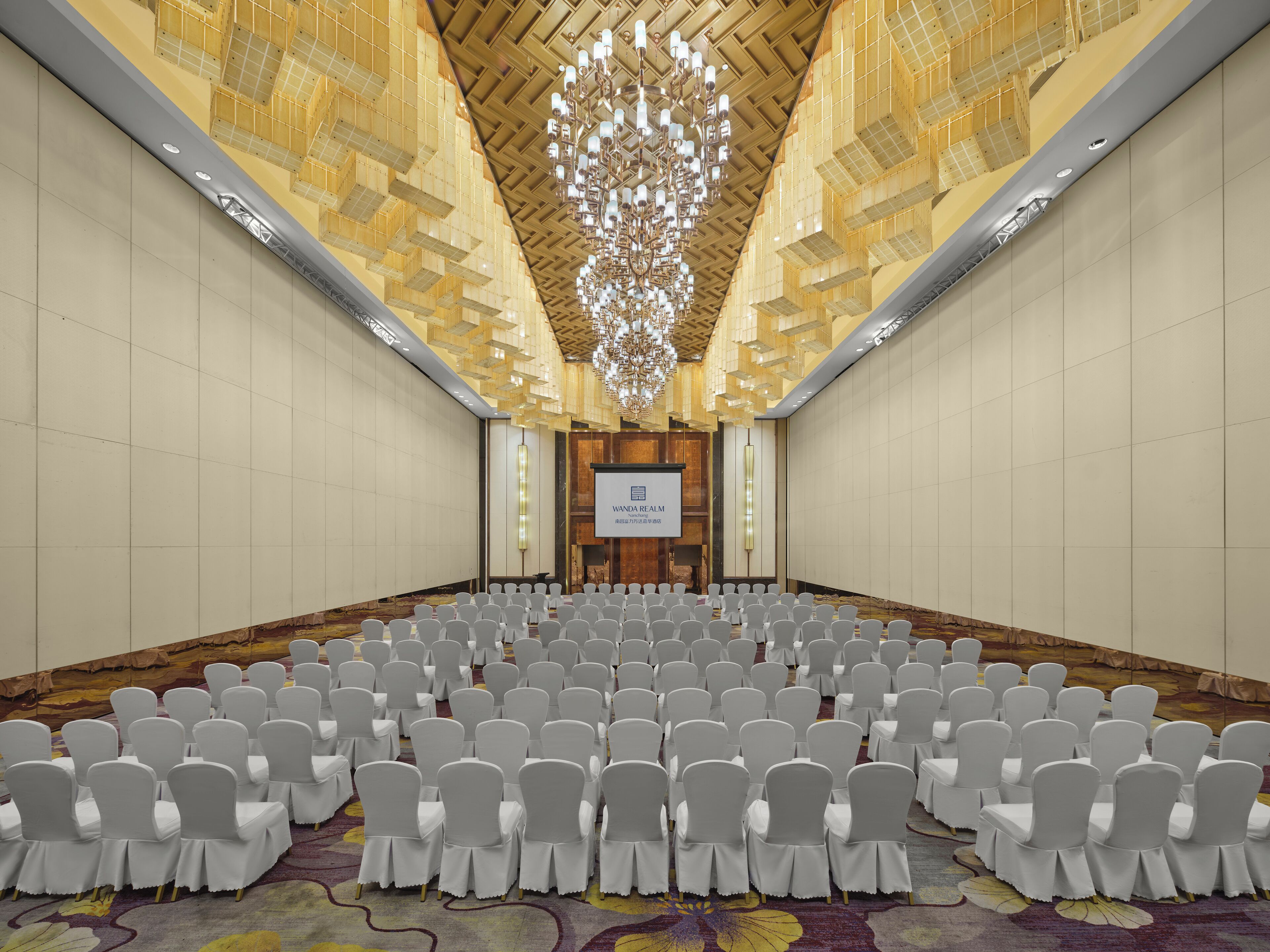 Un salón de conferencias con hileras de sillas blancas, una lámpara de araña grande y una pantalla mostrando 'VANDAL REALM'.