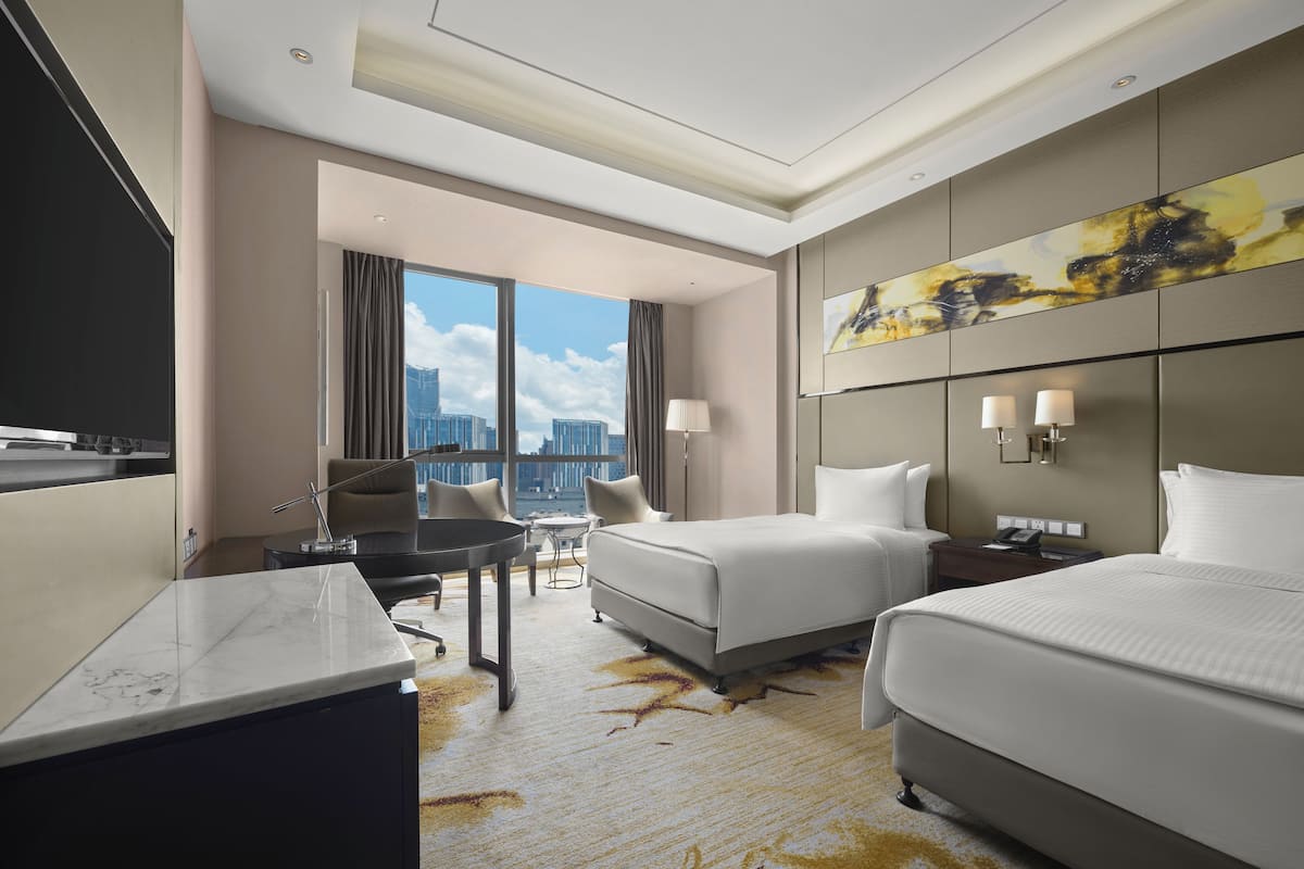 Ein Hotelzimmer mit zwei Betten, einem Schreibtisch, einem Stuhl und einem großen Fenster mit Blick auf die Stadt.