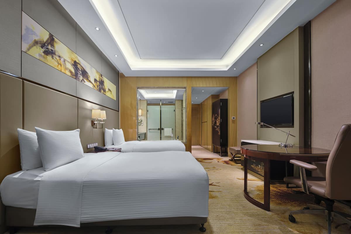 Ein modernes Hotelzimmer mit einem großen Bett, einem Schreibtisch, einem Stuhl, einem Fernseher und einem durch eine Glastür einsehbarer Badezimmer.