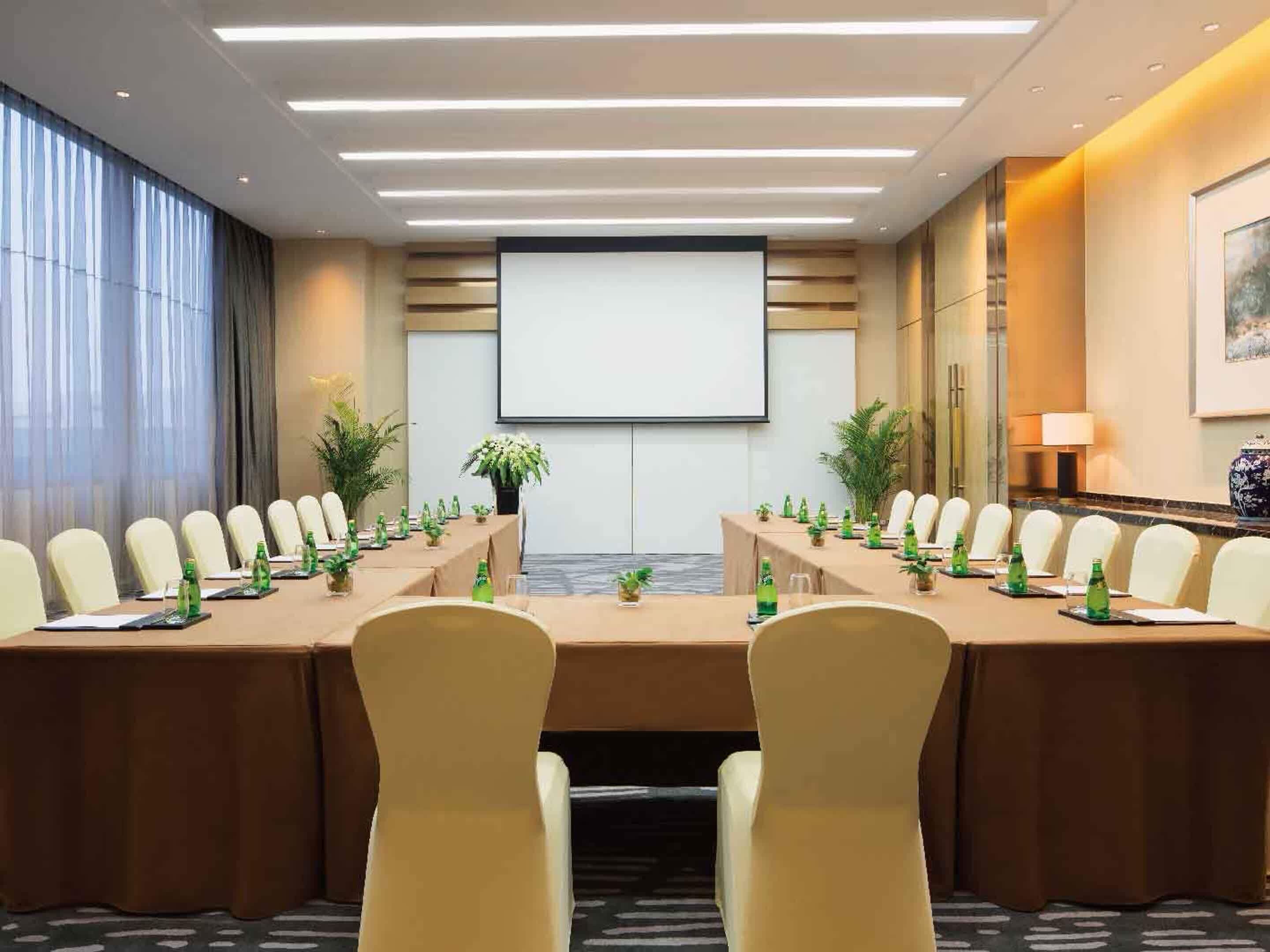 A conference room with a long table, chairs, a projector screen, and potted plants.