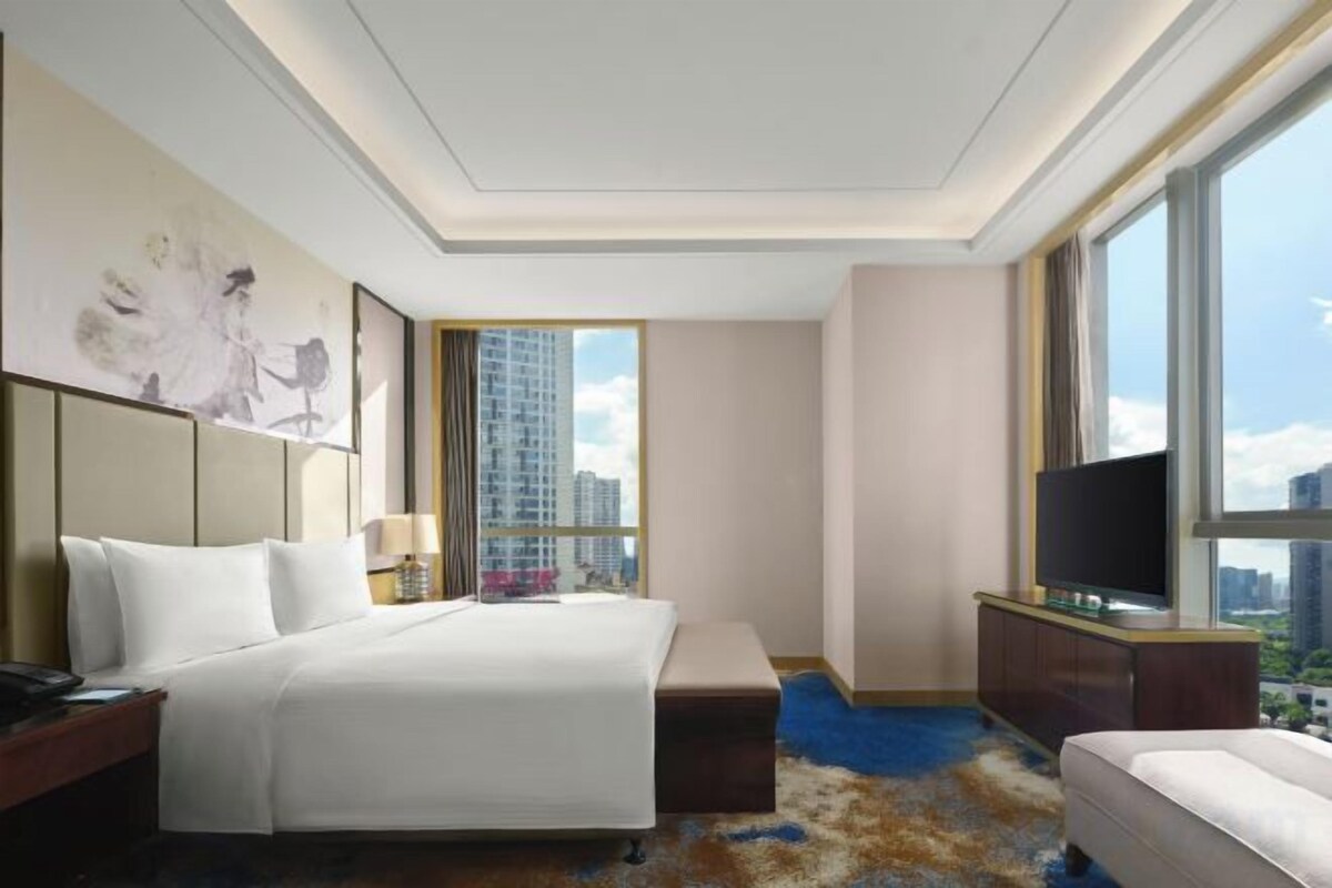 A hotel room with a large bed, a desk, a TV, and a view of the city.
