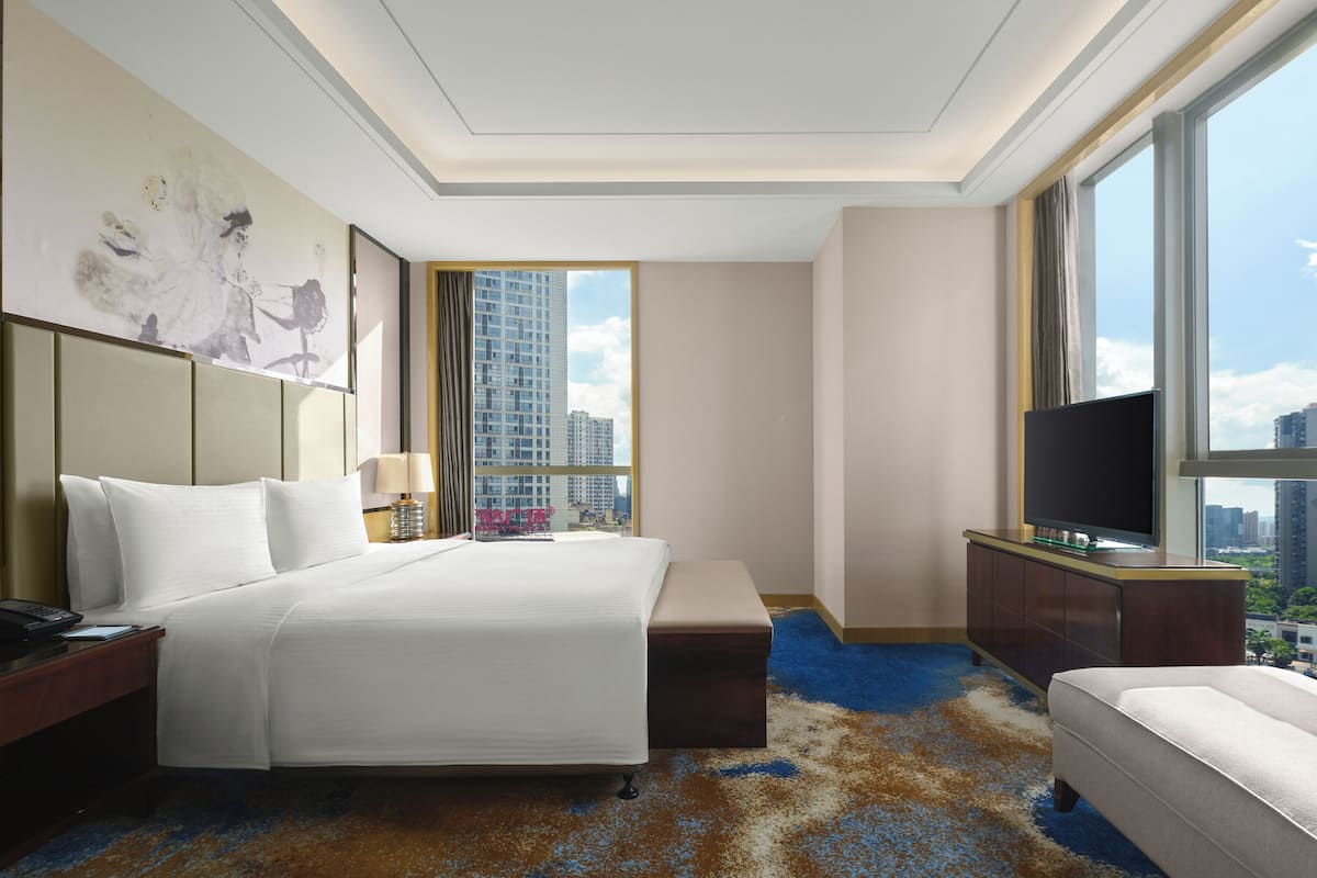 Ein Hotelzimmer mit einem großen Bett, einem Schreibtisch mit Lampe, einem Fernseher und Blick auf hohe Gebäude.