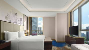 A hotel room with a large bed, a desk with a lamp, a TV, and a view of tall buildings.