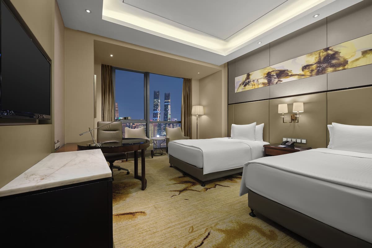 Ein Hotelzimmer mit zwei Betten, einem Schreibtisch, einem Sessel, einem Fernseher und einem großen Fenster mit Blick auf die Stadt.