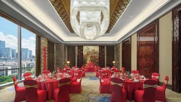Un salón de banquetes con mesas redondas dispuestas para un evento formal, grandes ventanales con vistas a la ciudad y una imponente araña.