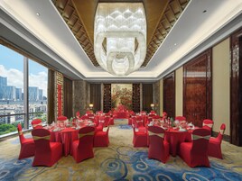 Un salón de banquetes con mesas redondas dispuestas para un evento formal, grandes ventanales con vistas a la ciudad y una imponente araña.