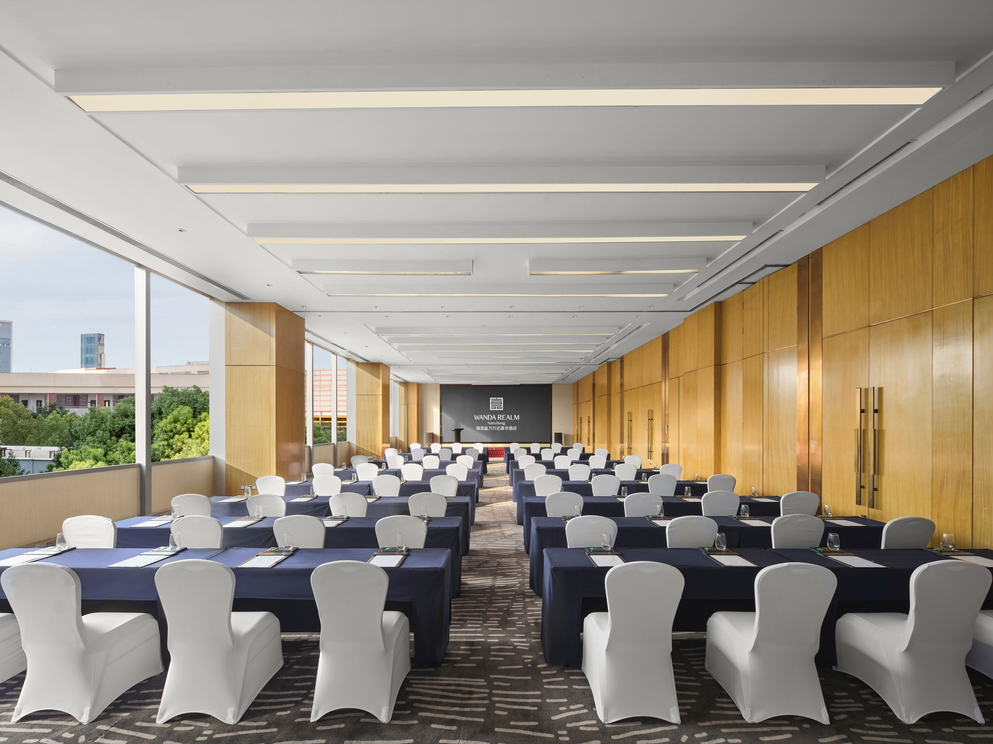 A conference room with rows of tables and chairs, a projector screen, and a view of the cityscape.