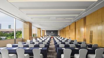 A conference room with rows of tables and chairs, a projector screen, and a view of the cityscape.