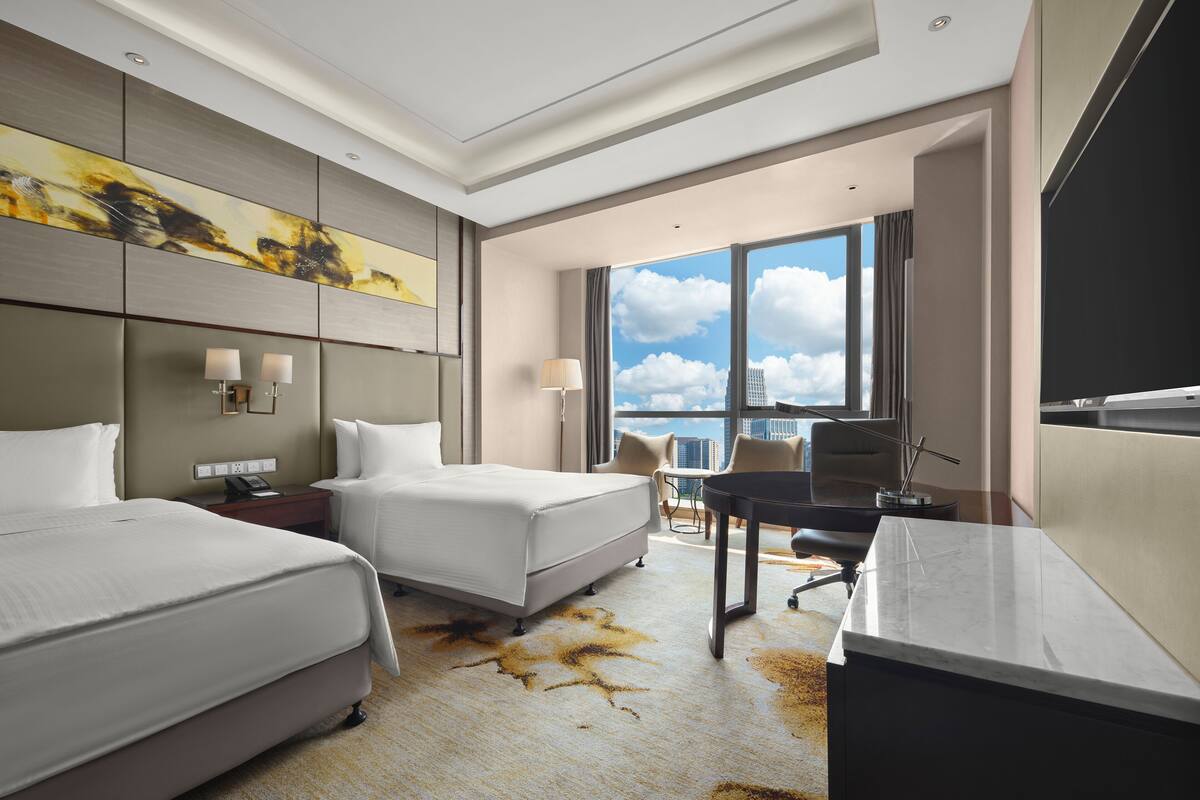 Ein Hotelzimmer mit zwei Betten, einem Schreibtisch, einem Stuhl, einer Lampe und einem großen Fenster mit Blick auf die Stadt.
