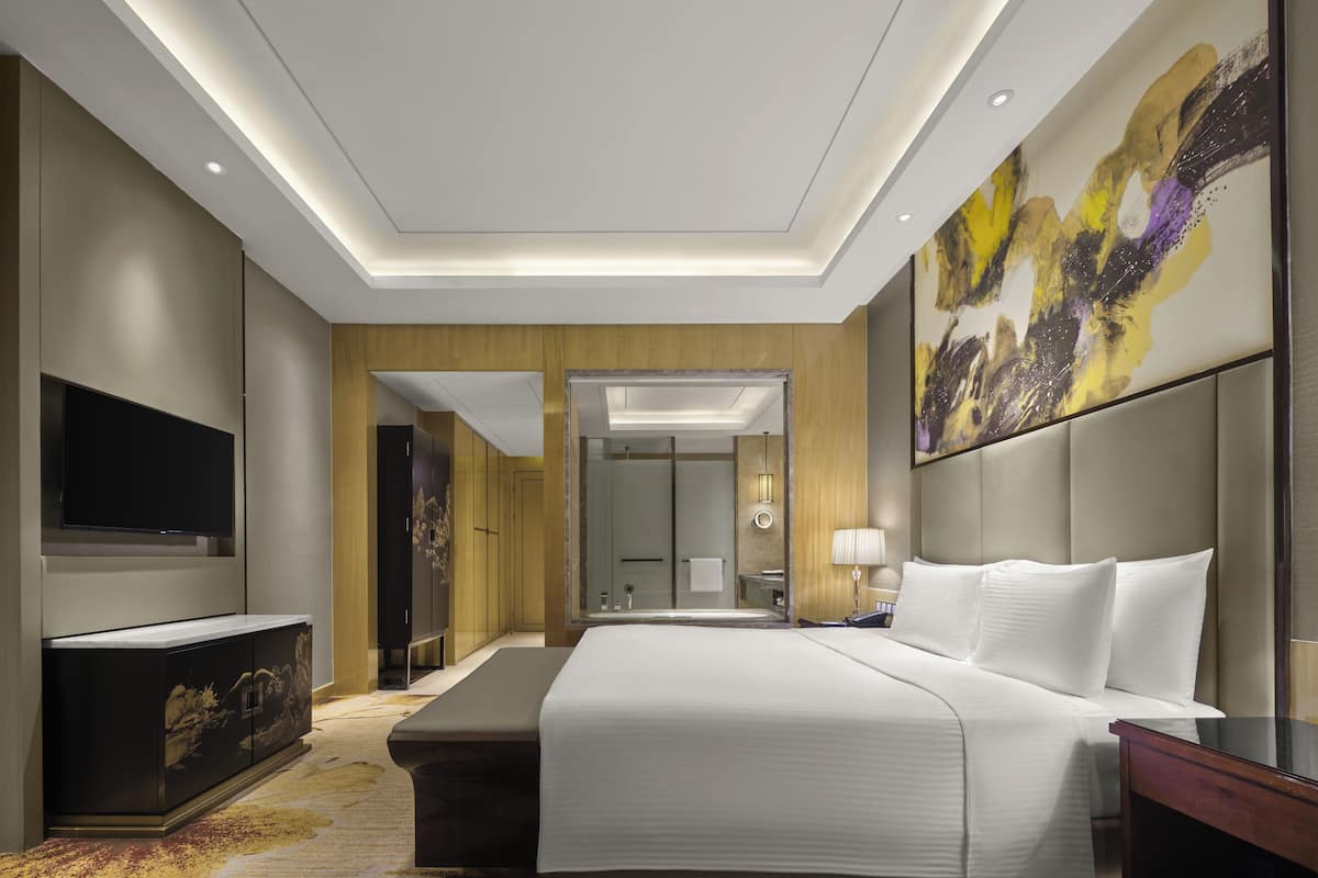 Ein modernes Hotelzimmer mit einem großen Bett, einem Flachbildfernseher, einem Schreibtisch und einem durchgegangene Tür sichtbaren Badezimmer.