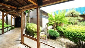 A wooden deck leading to a covered area with a view of a garden with various plants and a blue building in the background. - Hotel Ryusendo Aizan (Iwaizumi)