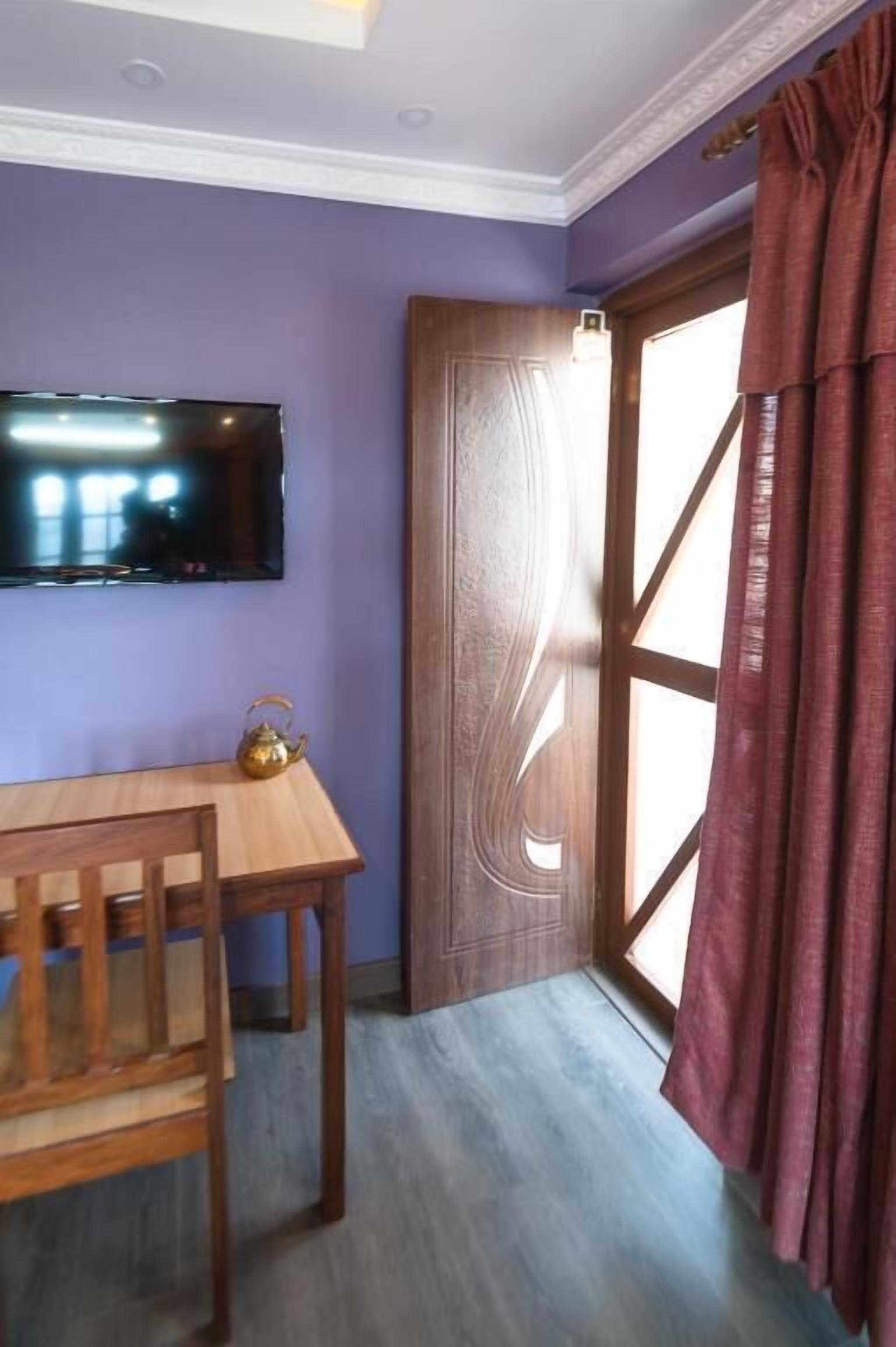 A room with a wooden table and chairs, a flat-screen TV mounted on the wall, and a large window with curtains.