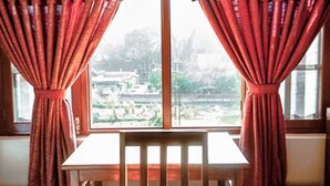 A room with a wooden table and bench, red curtains, and a window with a view of a landscape. - Be Here Now Guest House (Kathmandu)