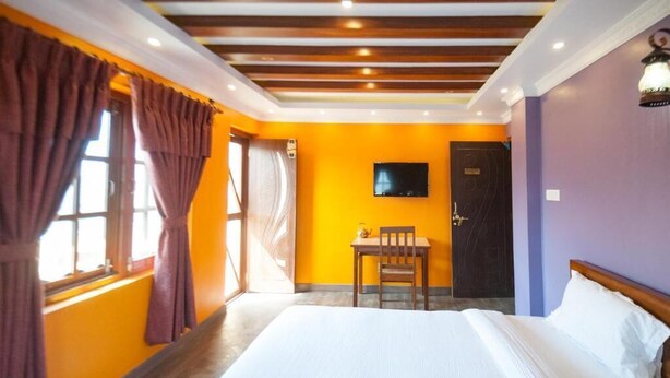 A hotel room with a large bed, a desk with a chair, a television on the wall, and a door. - Be Here Now Guest House (Kathmandu)
