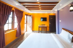 A hotel room with a large bed, a desk with a chair, a television on the wall, and a door. - Be Here Now Guest House (Kathmandu)