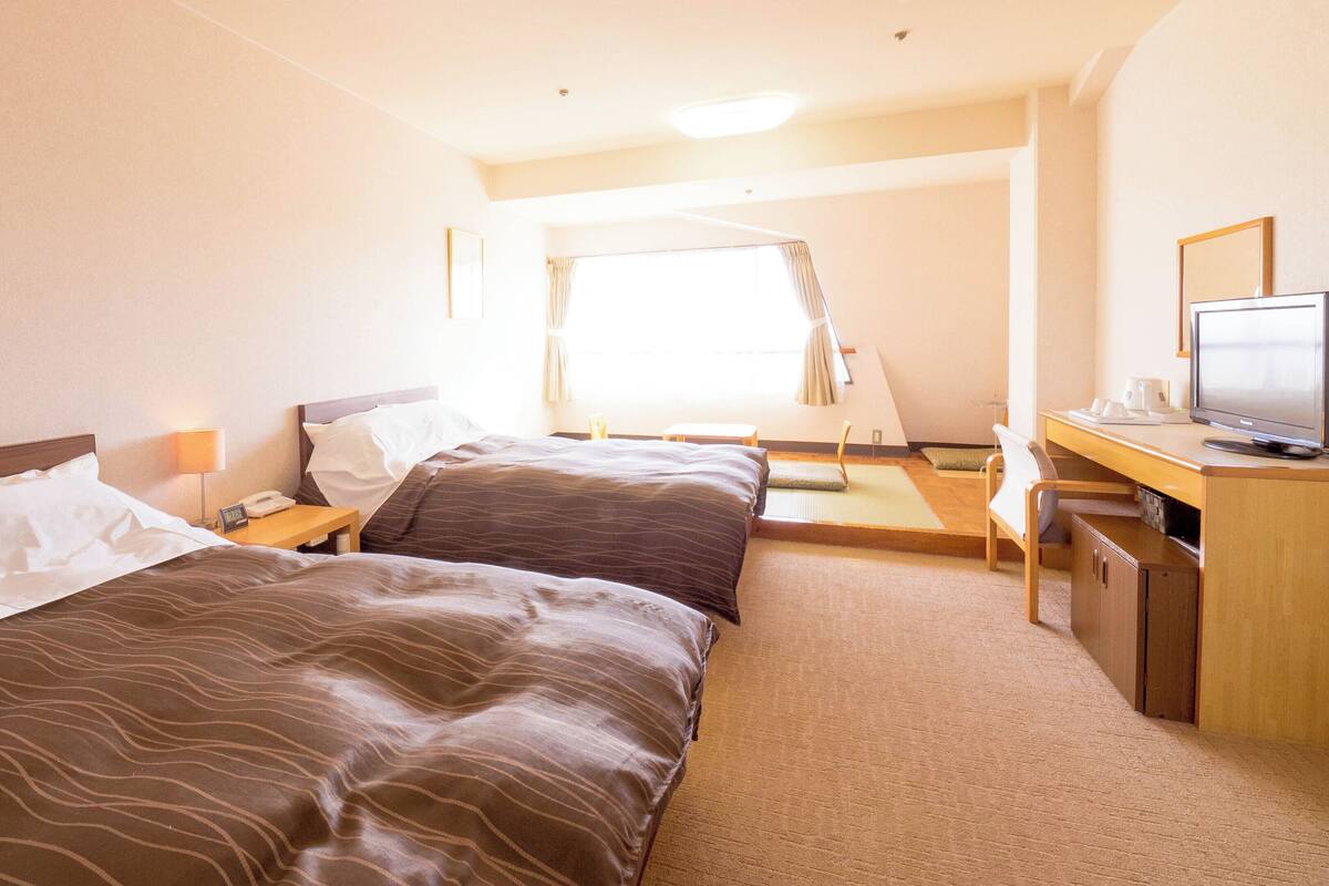 Quarto de hotel com duas camas, uma escrivaninha com televisão e uma janela com cortinas.