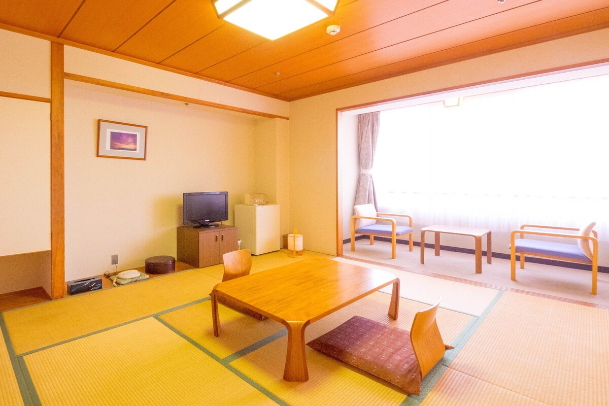 A traditional Japanese room with tatami flooring, a low table, a television, and a sliding door leading to a balcony.