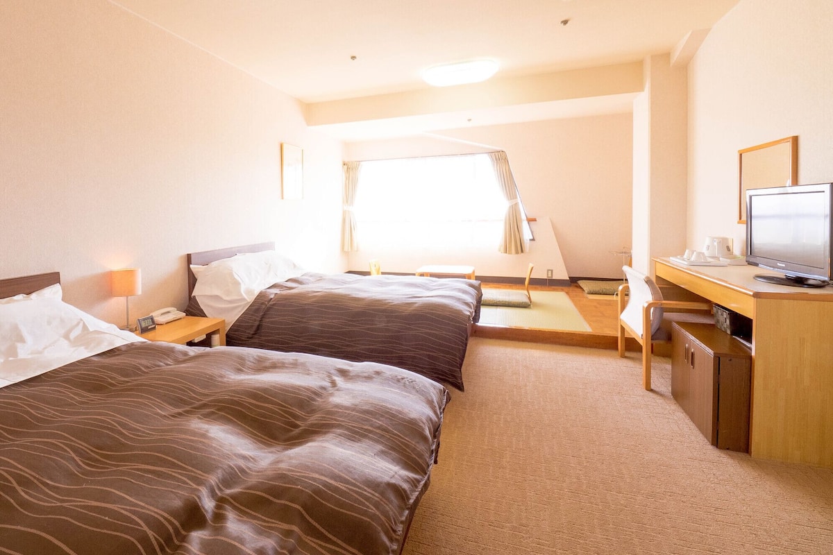 Quarto de hotel com duas camas, uma escrivaninha com televisão e uma janela com cortinas.