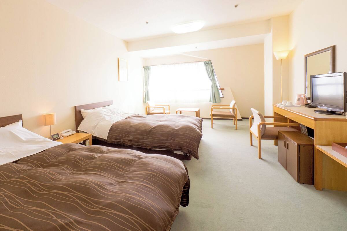 Quarto de hotel com duas camas, uma escrivaninha com computador, uma cadeira e uma janela com cortinas.