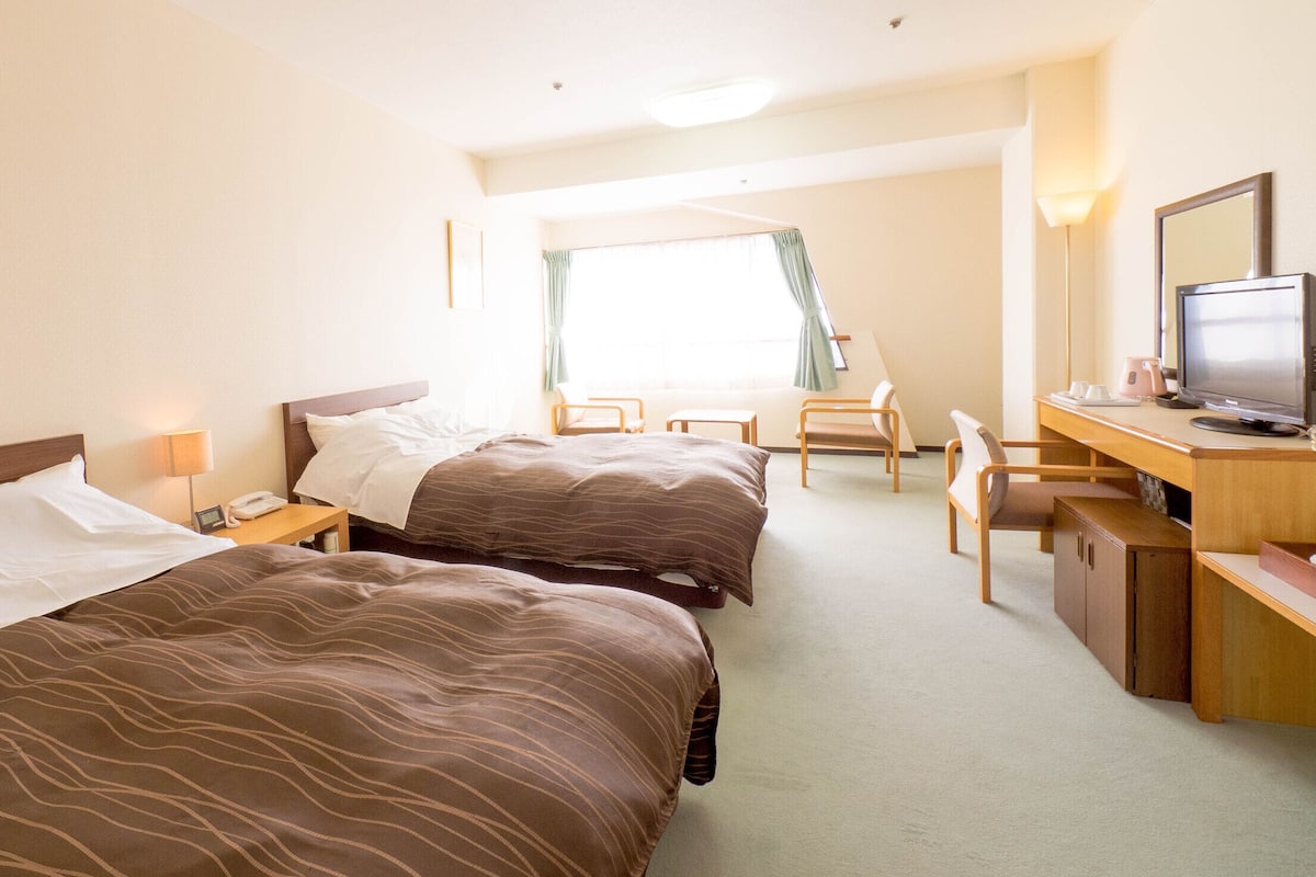 Quarto de hotel com duas camas, uma escrivaninha com computador, uma cadeira e uma janela com cortinas.
