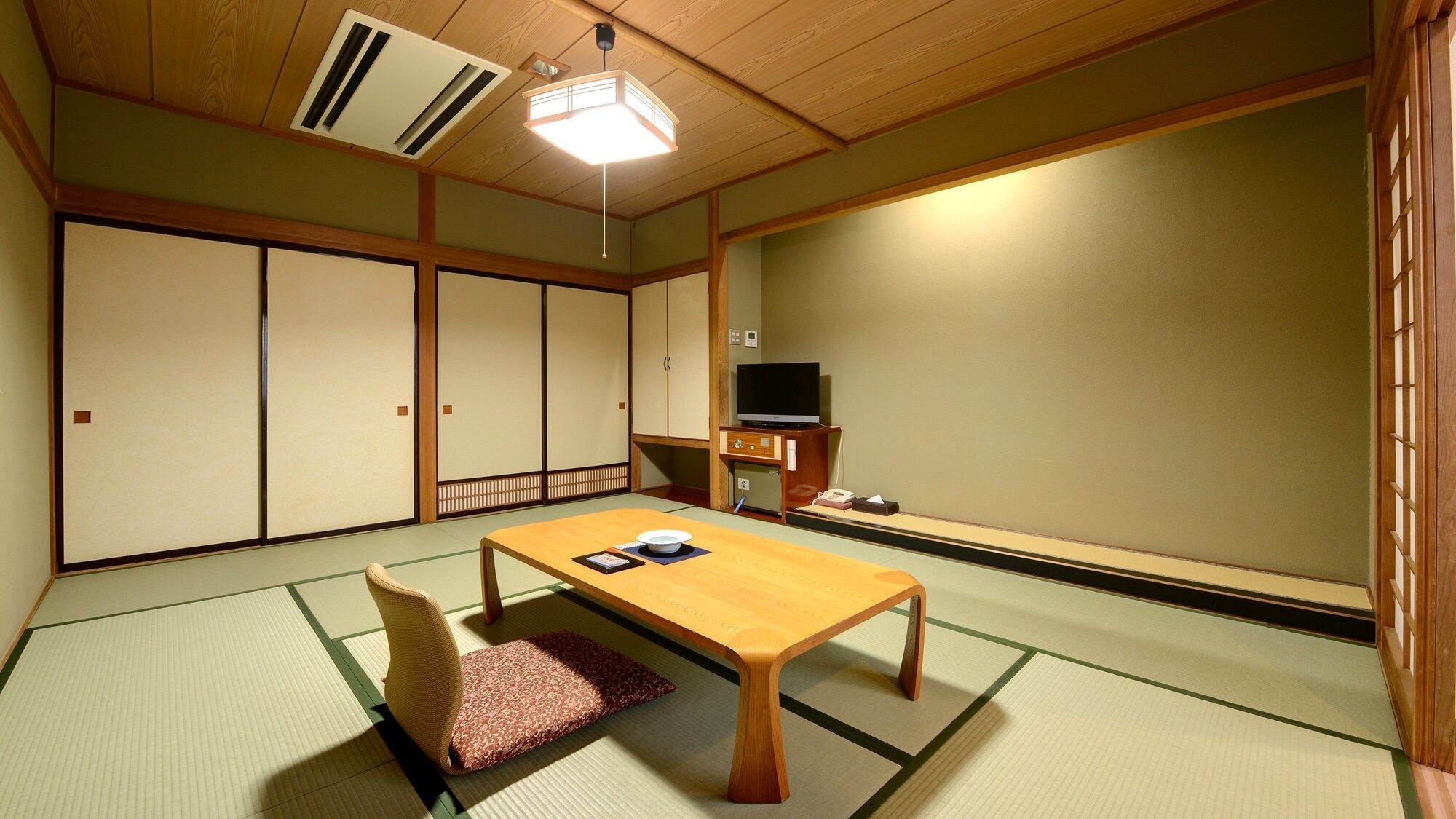 Una habitación japonesa tradicional con suelo de tatami, una mesa de madera baja, una silla acolchada, puertas correderas y un televisor.