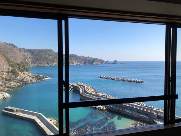 A coastal view through a window with a clear sky, rocky coastline, and a pier extending into the water. - Ryokan Hotel Ragaso (Tanohata)
