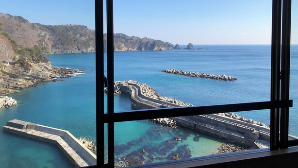 Une vue sur la côte, aperçue à travers une fenêtre, avec des falaises rocheuses, une mer calme et une jetée en pierre.