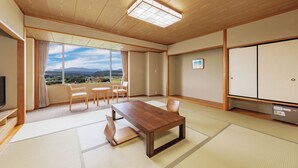 Una habitación japonesa tradicional con suelo de tatami, una mesa de madera, sillas, un televisor y vistas a las montañas.