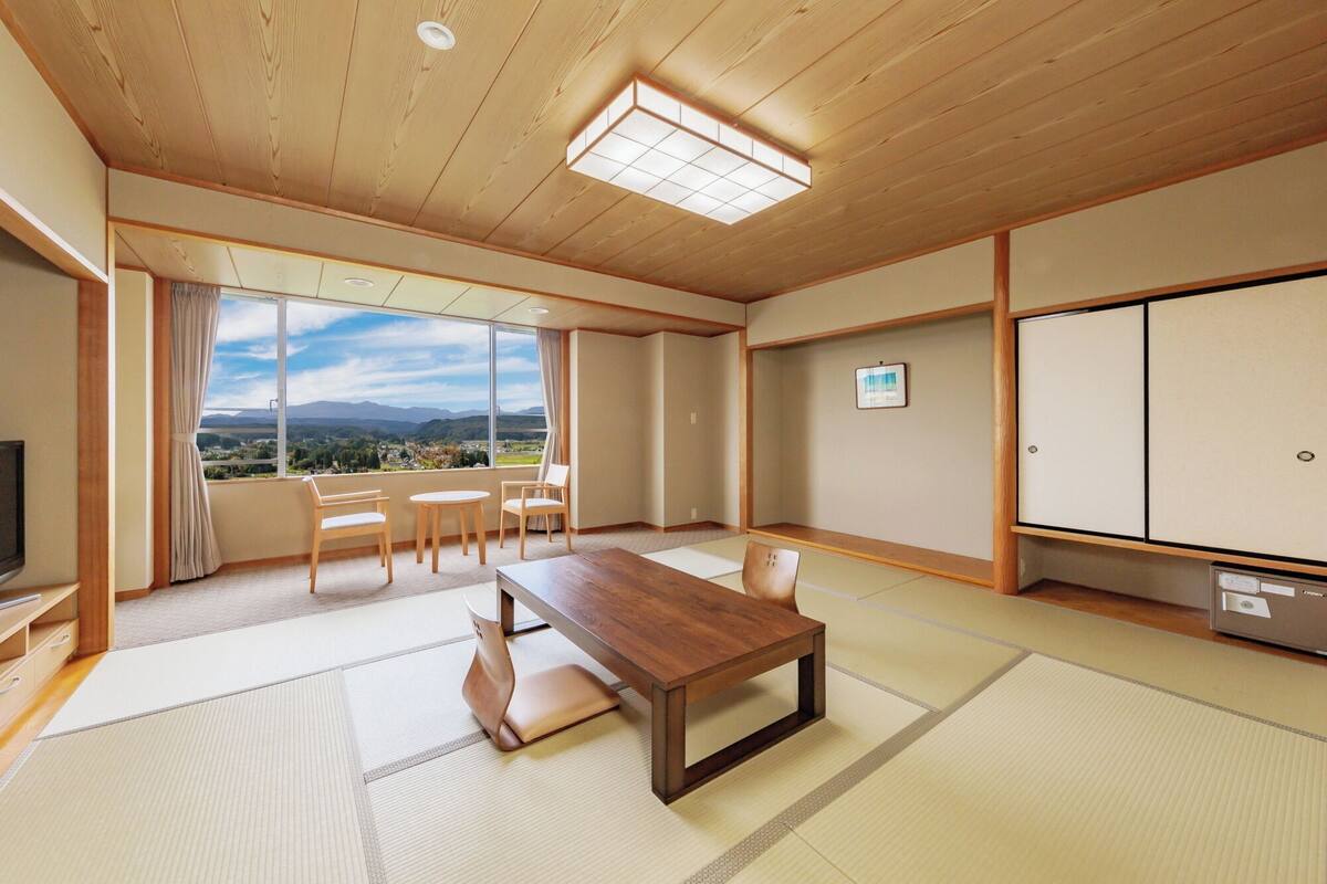 Una habitación japonesa tradicional con suelo de tatami, una mesa de madera, sillas, un televisor y vistas a las montañas.