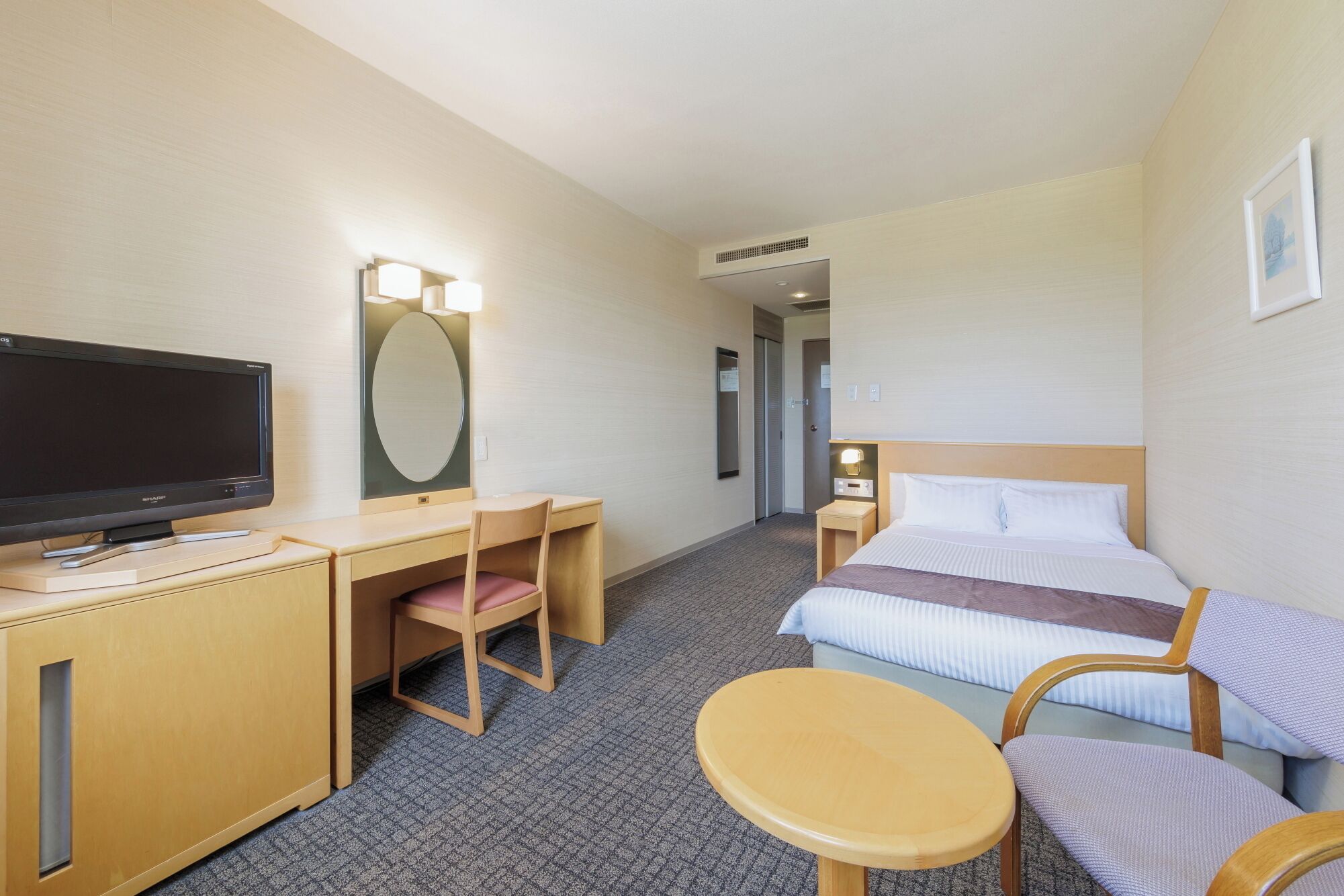 A hotel room with a bed, a desk with a television, a chair, a small table, and a mirror.