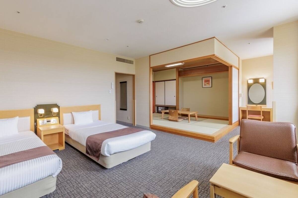 Habitación de hotel con dos camas, una zona de estar con una silla y una mesa, un espejo y una mesita de noche con lámpara.